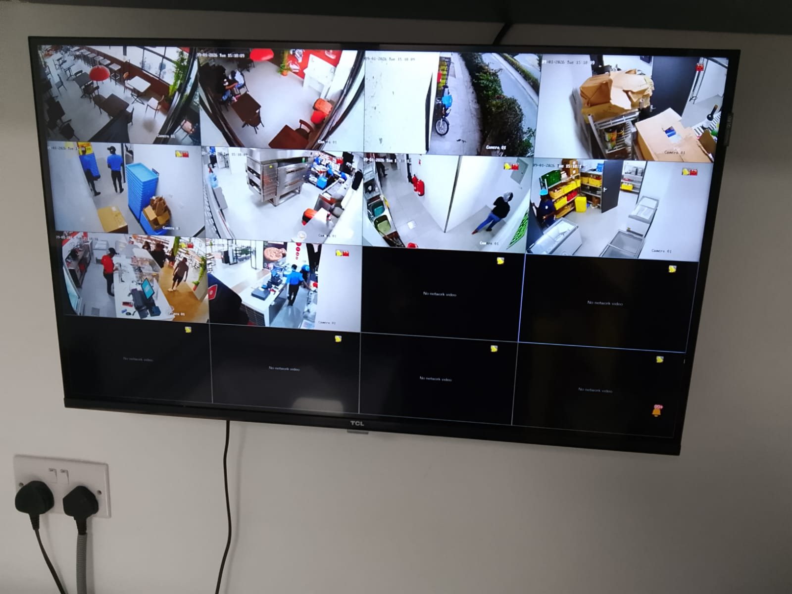 Wall-mounted CCTV monitor showing a live split-screen view of a restaurant dining area, kitchen, stores and entrance